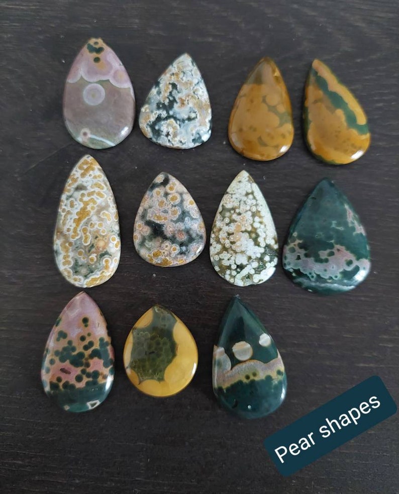 May include: Twelve pear-shaped ocean jasper stones in various shades of green, brown, and white with a speckled pattern. The stones are arranged in three rows of four on a dark brown surface. The text "Pear shapes" is written in white on a black background at the bottom of the image.