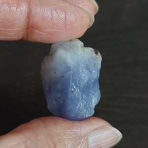 May include: A rough, light blue and white gemstone with a slightly irregular shape. The gemstone has a few dark spots on its surface.