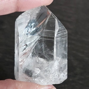May include: A clear quartz crystal point with a faceted shape and a natural, rough texture. The crystal is held in a hand, showcasing its size and clarity.