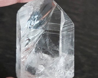 Puede incluir: Una punta de cristal de cuarzo transparente con forma facetada y textura natural rugosa. El cristal se sostiene en una mano, mostrando su tamaño y claridad.