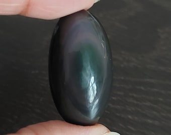 May include: A smooth, oval-shaped obsidian stone with a dark gray color and iridescent green and purple hues.