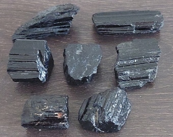 May include: Seven raw, black tourmaline crystals are arranged on a wooden surface. The crystals vary in size and shape, with a deep, glossy black color. Some pieces have visible striations and natural textures.