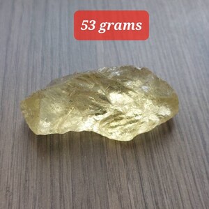K&ouml;nnte beinhalten: Ein gro&szlig;er, durchscheinender, gelb-goldener Kristall mit einem Gewicht von 53 Gramm. Der Kristall hat eine raue, nat&uuml;rliche Textur und liegt auf einer Holzoberfl&auml;che.