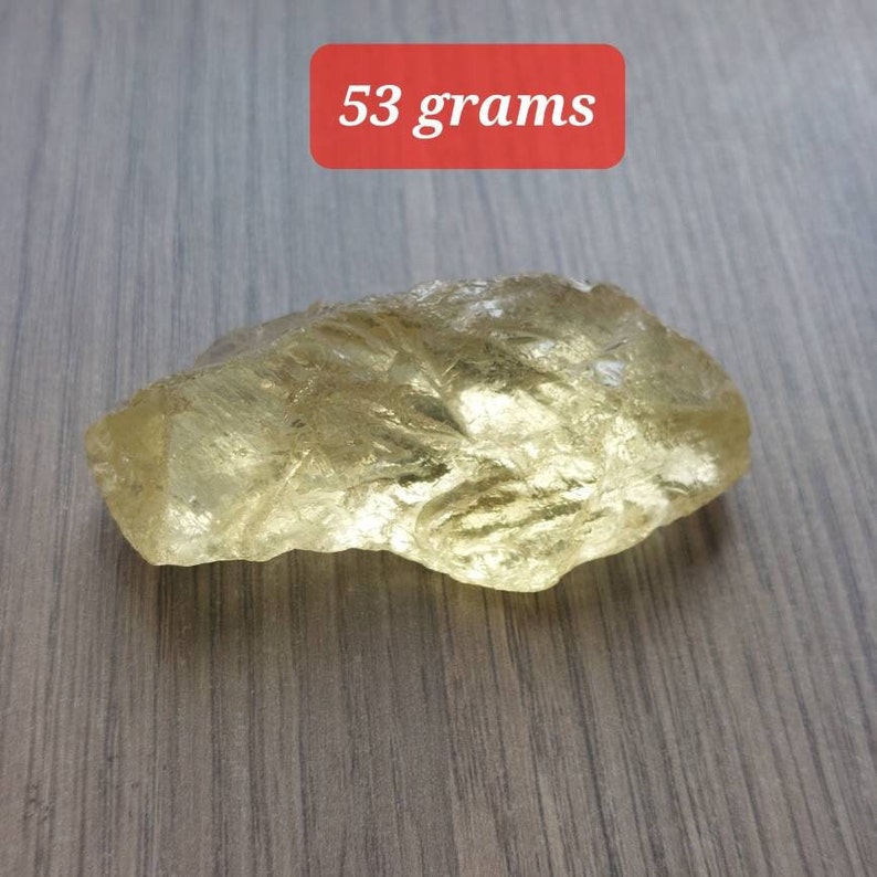 K&ouml;nnte beinhalten: Ein gro&szlig;er, durchscheinender, gelb-goldener Kristall mit einem Gewicht von 53 Gramm. Der Kristall hat eine raue, nat&uuml;rliche Textur und liegt auf einer Holzoberfl&auml;che.