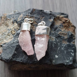 May include: Two pink quartz crystal pendants with silver settings. The crystals are raw and have a natural, irregular shape. The pendants are displayed on a dark gray rock.