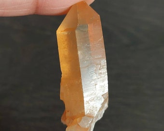 May include: A clear, orange crystal point with a pointed tip and a natural, rough base. The crystal is translucent and has a slight orange hue.