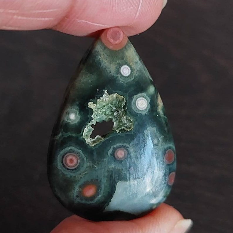 May include: A teardrop-shaped green, blue, and white ocean jasper pendant with red and pink spots. The pendant has a hole drilled through the top for stringing.