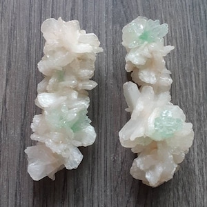 May include: Two clusters of white and pale green crystals. The crystals have a delicate, layered appearance, with a soft, almost floral structure. The color palette is primarily white with subtle hints of green, creating a natural and organic aesthetic.