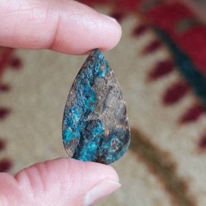 May include: A teardrop-shaped cabochon gemstone with a copper-toned base and blue-green inclusions. The stone has a rough, textured surface.