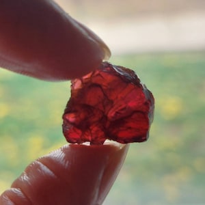 Puede incluir: Una piedra preciosa de corte tosco de color rojo intenso con una forma natural irregular. La piedra preciosa se sostiene entre dos dedos.