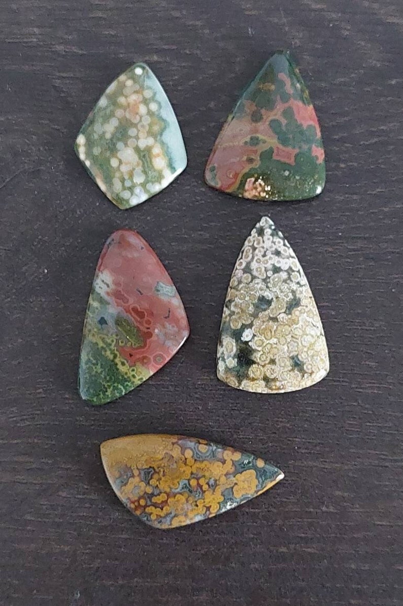 May include: Five irregularly shaped, polished, green, brown, and white ocean jasper stones. The stones have a variety of patterns and colors, including white spots, brown swirls, and green veins.