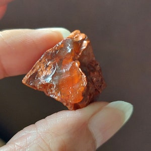 Op de afbeelding: Een ruwe, oranje-bruine edelsteen met een doorschijnend uiterlijk. De natuursteen heeft een grillige, onregelmatige vorm en een glanzend oppervlak. De edelsteen wordt tussen twee vingers gehouden tegen een vage bruine achtergrond.