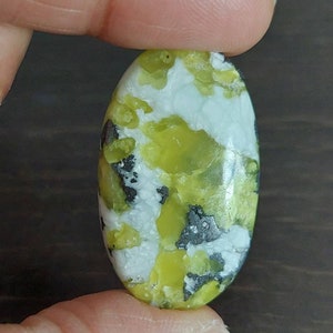 May include: Oval shaped green, white, and black gemstone cabochon. The stone has a mottled pattern with green, white, and black swirls.