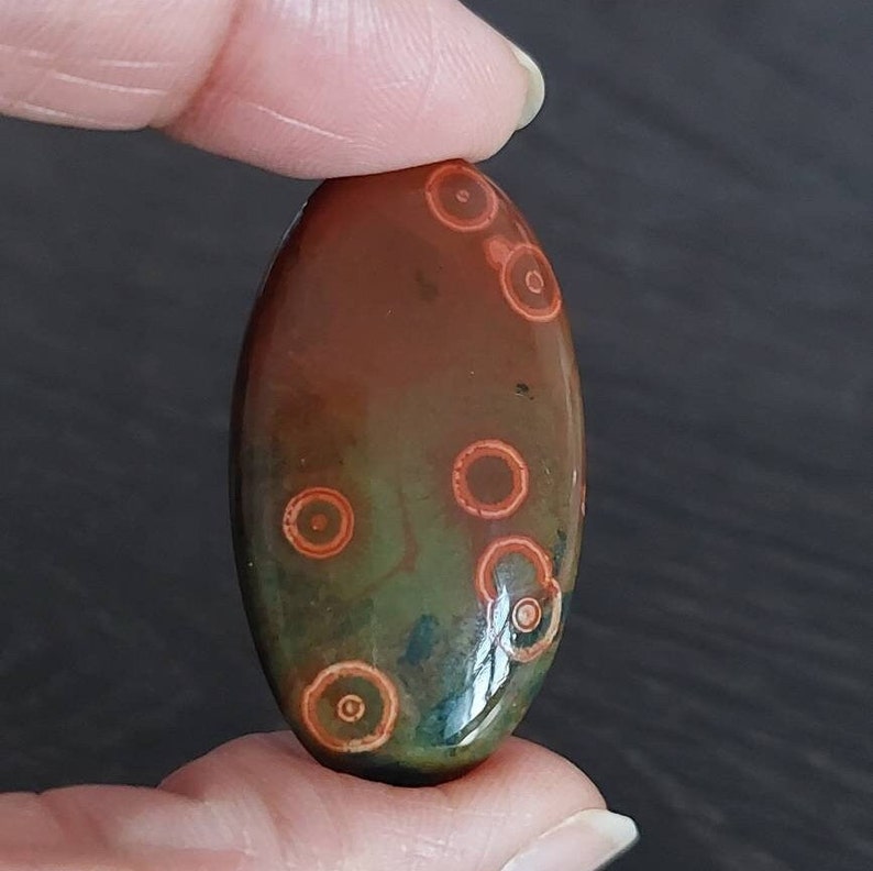 May include: A polished oval-shaped stone with a green and brown base and red and orange concentric circles.