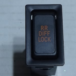 Puede incluir: Un interruptor automotriz negro con el texto "RR DIFF LOCK" en naranja. El interruptor es rectangular con un botón empotrado, probablemente para un bloqueo del diferencial trasero.