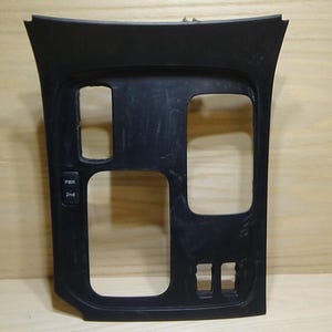 May include: A black plastic car dashboard trim piece with various openings for controls and displays. It has rectangular and square cutouts, and small buttons labeled "PWR" and "2nd". The trim is against a light brown wooden background.