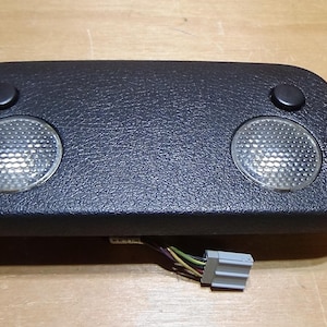 May include: Black overhead console featuring two dome lights and two small buttons. The console has a textured surface and a wire harness with a grey connector. The lights have a clear, textured lens.
