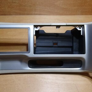 May include: A silver and beige automotive dashboard centre console trim panel featuring a rectangular opening for a radio or navigation unit, a secondary storage compartment, and a horizontal slot, constructed from moulded plastic components.
