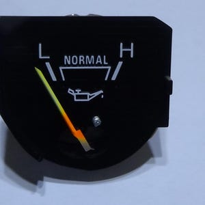 May include: Black oil pressure gauge with a yellow and orange needle. The gauge reads "L", "NORMAL", and "H". An oil can symbol is in the center. The gauge is set against a light background.