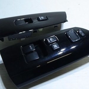 May include: Two black plastic car window switch panels. The top panel has a single window switch and the bottom panel has four switches, including an "AUTO" switch.