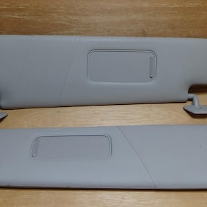 Op de afbeelding: Twee lichtgrijze zonnekleppen voor auto's. Elke zonneklep heeft een rechthoekige uitsparing en een draaiarm voor afstelling. De zonnekleppen zijn ontworpen om zonlicht te blokkeren en verblinding te verminderen.