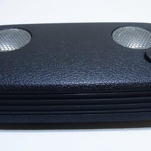 May include: A black, rectangular overhead console with two circular lights and two small buttons. The console has a textured surface and a ribbed lower edge. The lights have a mesh-like pattern.