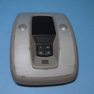 May include: A white overhead interior car light with a black control panel featuring buttons for light intensity and a small digital display. The light has two small white dome lights on either side of the control panel.