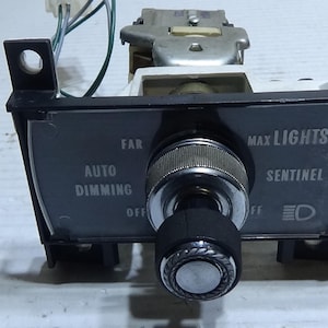 May include: A black plastic automotive headlight switch with a silver knob. The switch has labels for "Auto Dimming", "Far", "Max Lights", and "Sentinel".