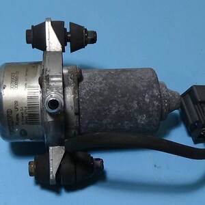 Op de afbeelding: Een zilveren autovacuümpomp met een zwarte elektrische connector en een zwarte draad. De pomp heeft een label met de tekst "03C129800 EX2001".