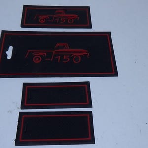 May include: A set of black rubber mats with red trim. The mats feature a red outline of a vintage pickup truck and the number "150". The set includes a large mat, two smaller rectangular mats, and two other mats.