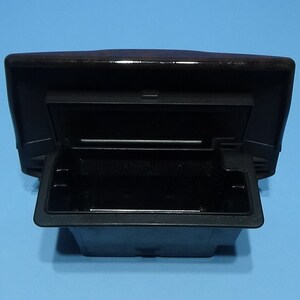 May include: A black, rectangular car ashtray with an open lid. The ashtray is made of plastic and has a compartment for ashes and a lid that opens and closes. The background is a solid light blue.