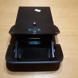 May include: A black electronic device with a hinged top and an open compartment. The device has a small sticker on the top and a series of vertical lines inside the open compartment. The device is on a light brown wooden surface.