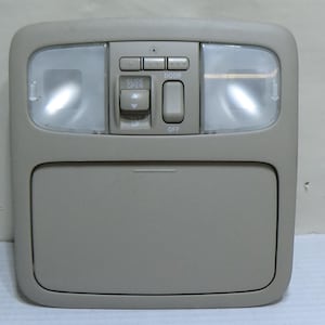 May include: Tan-colored overhead console with integrated lights and control buttons. Features two bright lights, a storage compartment, and several buttons for various functions. The console is designed for automotive use.