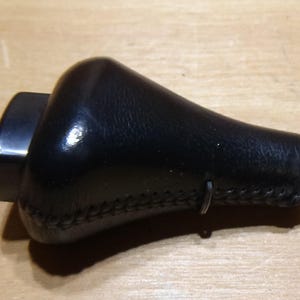 Peut inclure: Un pommeau de levier de vitesses en cuir noir avec un dessus arrondi et une base cylindrique. Le pommeau a une couture cousue autour du bas et un clip métallique. L'objet est posé sur une surface en bois brun clair.