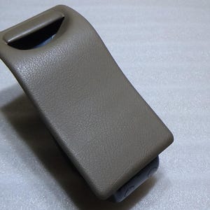 May include: A tan-colored, curved plastic component with a textured surface. It features a recessed handle at the top and a rectangular base. The item appears to be a car interior part.