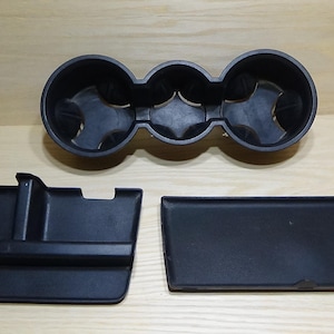 Könnte beinhalten: Schwarze Kunststoff-Autokonsolenkomponenten, darunter ein dreifacher Getränkehalter, eine rechteckige Ablage und ein Fach mit erhöhtem Rand. Die Artikel sind auf einer hellen Holzoberfläche angeordnet.