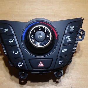 May include: Black car climate control panel with a central dial for temperature adjustment, marked with numbers and a blue-to-red color gradient. Features buttons for fan speed, air direction, and A/C, plus a hazard light button.