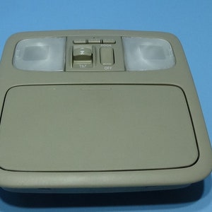 May include: A beige car interior dome light with two reading lights and a control panel with buttons labeled "Tilt" and "Off".