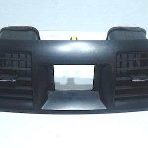 Puede incluir: Rejilla de ventilación de coche de plástico negro con dos aberturas rectangulares y un panel central rectangular. La rejilla tiene una superficie texturizada y está diseñada para encajar en el salpicadero de un coche.
