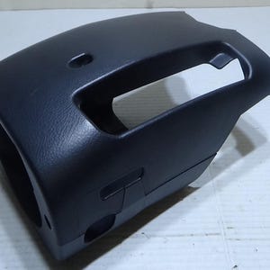 May include: A black plastic steering column cover with a textured surface. The cover has a large rectangular opening and a smaller circular opening. It is designed to fit around the steering column.
