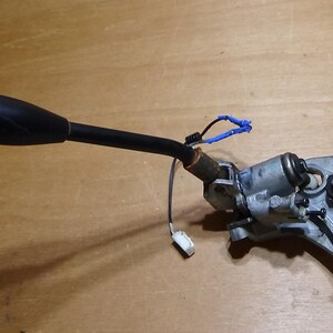 May include: A black automotive gear shift lever with a metallic silver mounting bracket and base. The assembly includes a thin black wire with a small white plastic connector and a short blue wire segment attached to the central metal pivot point.
