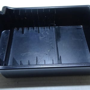 Puede incluir: Un contenedor de almacenamiento de plástico negro con forma rectangular. El contenedor tiene una serie de divisores verticales y una base plana. El contenedor tiene una pequeña pestaña en el lateral. El contenedor está vacío.