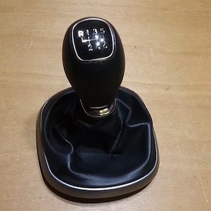 Peut inclure: Un pommeau de levier de vitesses noir avec un dessus bordé de chrome, affichant les positions des vitesses. Le pommeau est monté sur un soufflet en similicuir noir avec une bordure argentée. Fond en bois brun clair.