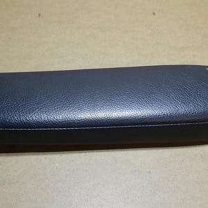May include: A dark blue, rectangular automotive interior component with a textured surface. The piece has a rounded end and a stitched seam along the edge. The item appears to be made of a synthetic material.