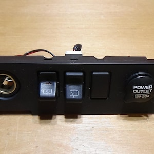 May include: Black automotive control panel featuring a cigarette lighter, three switches, and a power outlet marked "POWER OUTLET 12V-20A". The panel is rectangular with mounting tabs on each end.