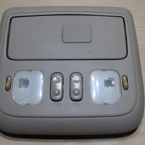 Puede incluir: Una consola de techo gris con un compartimento de almacenamiento y tres botones de control. La consola tiene dos luces cuadradas y dos pequeñas luces redondeadas. La consola es de plástico.