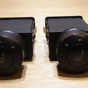 Puede incluir: Dos interruptores negros de control de calefacción de asientos de coche. Cada interruptor tiene un dial redondo negro con un borde texturizado y un símbolo de calefacción de asiento. Los interruptores son rectangulares y tienen clips metálicos.