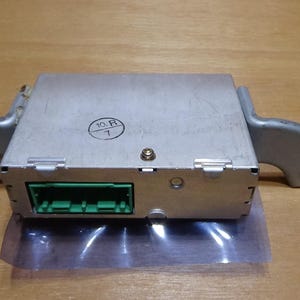 May include: A silver-colored electronic control module with a green connector and mounting brackets. The module has a small circular sticker with the numbers "10. R" and "7". The module is resting on a clear plastic sheet.