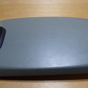 May include: A grey, rectangular automotive armrest with a black, curved handle on the left side. The armrest is made of a smooth, solid material and is designed to fit in a vehicle's interior.