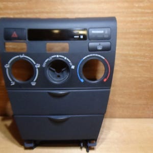 Puede incluir: Un panel de consola central de salpicadero de coche gris oscuro con tres aberturas circulares para mandos de climatización, una zona de pantalla rectangular, un botón de luces de emergencia y un botón de desempañador trasero.